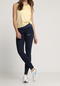 Top sin mangas de Nike amarillo con un gran logo, combinado con leggings navy y zapatillas deportivas blancas con acentos azules. Tejido suave, diseño ajustado.