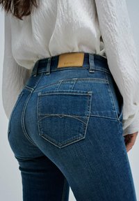 Mujer vistiendo jeans ajustados de mezclilla azul y una blusa blanca holgada, mostrada desde atrás a la altura de la cintura y las caderas.