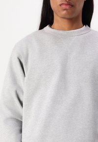 Šedá mikina s klasickým kulatým výstřihem a hladkým, strukturálním materiálem. Má ribovaný límec a švy přes ramena.