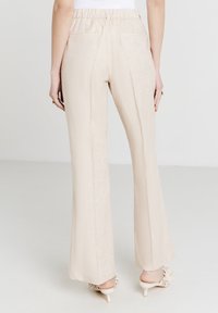Donna che indossa pantaloni larghi beige con elastico in vita e mule con tacco color crema con dettagli floreali, mostrata da dietro su sfondo neutro.
