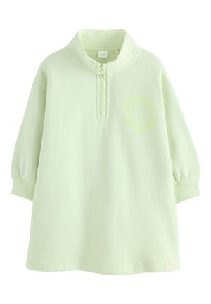 Enfant souriant debout dans une robe zippée vert clair et des baskets blanches montantes avec des chaussettes roses, fond neutre et objet circulaire texturé à proximité.