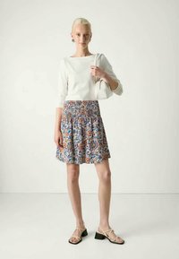 Fehér, hosszú ujjú felső, amely egy színes virágmintás szoknyával van párosítva, smockolt derékkal. A modellen bézs szandálok találhatók.