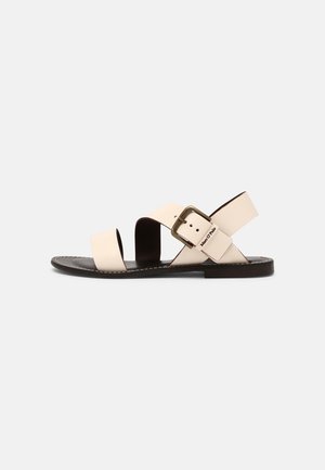 Sandalen met een platte zwarte zool, beige leren banden die over de voet korsen, en een rechthoekige metalen gesp voor aanpassing.
