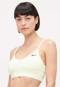 Soutien-gorge de sport vert clair avec un design en V, des bretelles fines et un petit logo Nike noir sur le côté gauche. Tissu lisse et extensible.