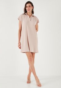 Robe polo rose pâle à manches courtes, coupe décontractée et décolleté en V. Tissu en mélange de coton doux, longueur genoux, associée à des sandales à lanières.