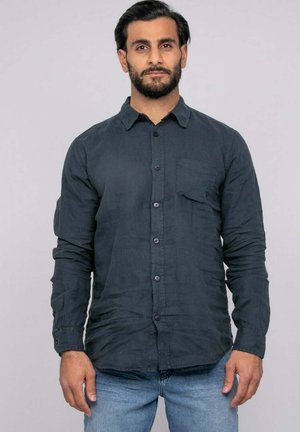 Homme aux cheveux foncés et à la barbe portant une chemise gris foncé à boutons et un jean bleu clair sur un fond clair uni.
