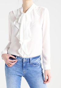 Blusa bianca a maniche lunghe con frontale arricciato e colletto a fiocco, abbinata a jeans azzurri chiari. Tessuto morbido, texture liscia e design aderente.