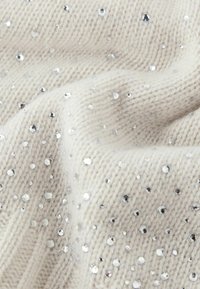 Kremowa dzianina z rozproszonymi srebrnymi cekinami, prezentująca miękką fakturę i lekko wypukły wzór dzięki cekinom.