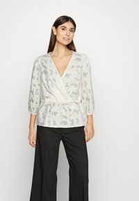 Blusa envolvente leve e texturizada em creme com padrões abstratos em cinza, apresentando um decote em V e detalhe de laço na cintura. Mangas longas e bufantes.