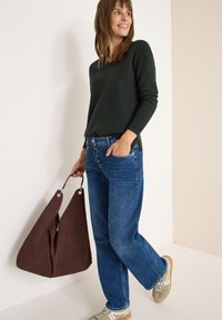 Dunkelgrüner Pullover, hoch geschnittene blaue Jeans und braune Wildleder-Handtasche. Hellfarbene Sneakers mit weißen Akzenten und strukturierter Oberfläche.