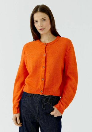 Frau trägt eine hell orangefarbene, geknöpfte Strickjacke und dunkelblaue Jeans, steht mit einer Hand in der Tasche vor einem schlichten hellen Hintergrund.