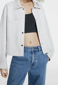 Veste en denim gris clair avec un col et des boutons argentés, portée sur un dessus côtelé foncé et un jean bleu taille haute. Comprend deux poches avant.