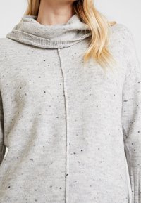 Maglione grigio chiaro con collo a cowl, lavorazione a maglia testurizzata e motivo a macchie in grigio scuro. Presenta una cucitura centrale e maniche lunghe.
