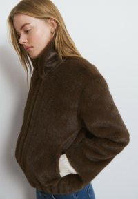 Chaqueta de piel sintética marrón con cuello alto, que presenta una textura suave, bolsillos laterales y puños de punto en color crema contrastante. Forma clásica y cuadrada.