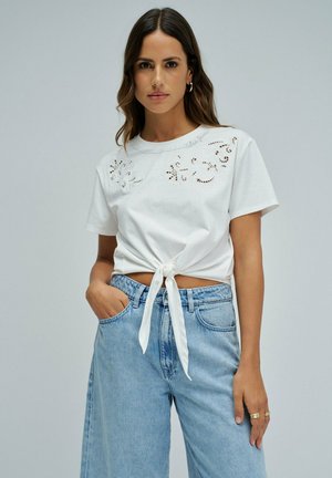 Femme aux cheveux bruns mi-longs porte un haut blanc à manches courtes avec des détails en dentelle noué devant et un jean bleu clair taille haute.