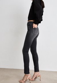 Vaqueros negros ajustados con una textura suave, cintura alta y diseño clásico de cinco bolsillos. Combinados con sandalias de tacón nude y una sudadera corta negra.