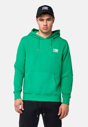 Homme portant un sweat à capuche vert avec une poche avant et une casquette noire, tous deux étiquetés "LEONE 1947,", debout devant un fond clair et uni.
