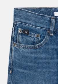 Jeans in denim blu con effetto sbiadito chiaro, dotati di una piccola tasca frontale con il logo "cK" in bianco e nero e cuciture a contrasto di colore oro.