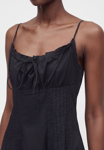 Gros plan d'une personne portant une robe noire sans manches avec des bretelles fines et un détail de nœud à l'avant au niveau du décolleté.