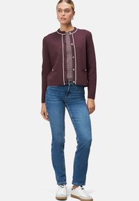 Woman wearing a burgundy buttoned cardigan over a striped shirt, blue jeans, and white sneakers, standing against a plain background.
