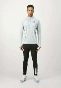 Jasnoszara, długorękawowa koszulka sportowa z zamkiem błyskawicznym na ćwierć, z logo na klatce piersiowej; zestawiona z czarnymi dopasowanymi spodniami i białymi butami sportowymi.