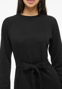 Žena v černém žebrovaném tričku s dlouhým rukávem, kulatým výstřihem a páskem ze stejné látky uvázaným na mašli v pase.