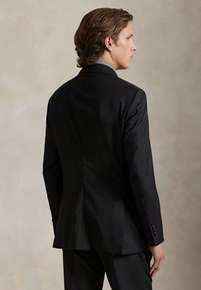 Mann mit mittellangen braunen Haaren, trägt eine schwarze Anzugsjacke und Hose, leicht zur Seite gedreht vor einem einfarbigen Hintergrund.