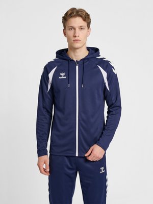 Młody mężczyzna ubrany w granatowo-białą sportową kurtkę na zamek oraz dopasowane spodnie z logo marki na piersi i udzie, stojący na jednolitym tle.