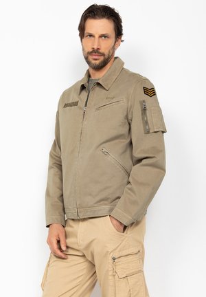 Mężczyzna z brodą, ubrany w beżową kurtkę w stylu wojskowym z naszywkami oraz beżowe spodnie typu cargo, stojący na tle białego tła.