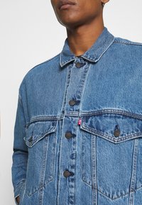 Denim jacket in medium blue, featuring a classic collar, two chest pockets, and brown snap buttons. Fabric has a textured finish.
