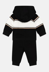 Ensemble en hoodie noir et pantalon de jogging avec des rayures horizontales blanches et marron, en matière coton et avec des poignets côtelés. Texture douce, design décontracté.