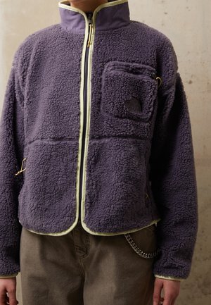 Osoba ubrana w fioletową polarową kurtkę z suwakiem z przodu, kieszenią na suwak na piersi i jasnym wykończeniem, na brązowych spodniach z dodatkiem łańcucha.