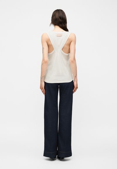 Γυναίκα με μακριά καστανά μαλλιά, που φοράει λευκό αμάνικο τοπ racerback και σκούρο φαρδύ παντελόνι, στέκεται με την πλάτη σε ένα απλό φόντο.