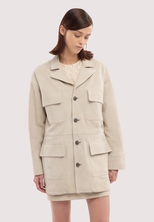 Frau trägt eine beige Jacke mit Knöpfen und vier aufgesetzten Taschen vorne über einem hellen, floralen Oberteil, steht vor einem schlichten Hintergrund.