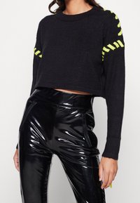 Svart kort tröja med neongula ränder på ärmarna, kombinerad med blanka svarta högmidjade byxor med dragkedjefickor.