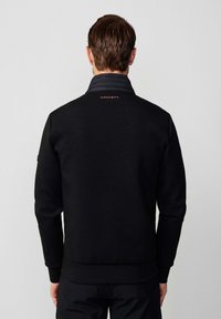 Homme aux cheveux bruns courts portant une veste noire avec le texte "Hackett" dans le haut du dos, debout devant un fond gris clair uni.