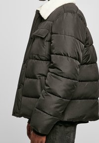 Puffjakke i mørkegrå med krave af kunstpels, med en firkantet form, quiltet tekstur og en lomme på venstre ærme.