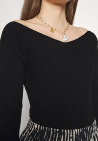 Top negro de manga larga con diseño acanalado y escote fuera del hombro, combinado con un collar de cadena dorada que presenta un colgante circular y un acento de perla.