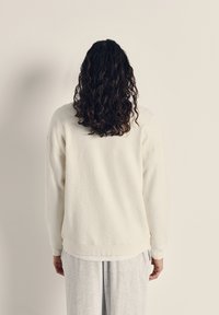 Vit fleece sweatshirt med en texturerad yta, ribbade ärmar och nederkant. Buren över ljusgrå sweatpants, sedd bakifrån.
