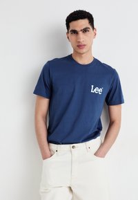 T-shirt en coton bleu foncé à manches courtes et col rond, avec un logo "Lee" blanc sur la poitrine gauche. Porté avec un pantalon de couleur claire.