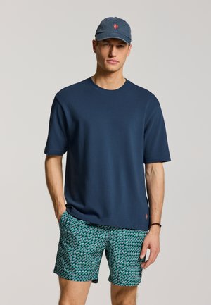 Man draagt een marineblauw T-shirt, groene shorts met patroon en een blauwe baseballpet met rode borduur, staat met één hand in zijn zak.