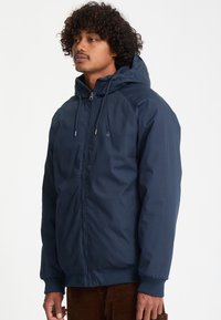Homme aux cheveux bouclés portant une veste à capuche bleu marine zippée et un pantalon marron, debout devant un fond clair uni.