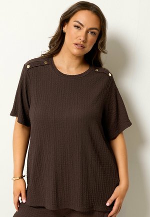 Femme portant un haut court texturé marron foncé avec des boutons dorés sur les épaules, debout devant un fond clair uni.