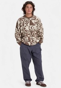 Νεαρός στέκεται με τα χέρια στις τσέπες, φορώντας έναν καφέ και κρεμ πουλόβερ με σχέδια, χαλαρούς σκούρους παντελόνες και καφέ αθλητικά παπούτσια με λευκές ρίγες.