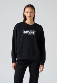 Felpa nera in cotone con colletto a girocollo, caratterizzata da un logo Levi's bianco a texture sul davanti. Il modello la indossa con jeans neri.