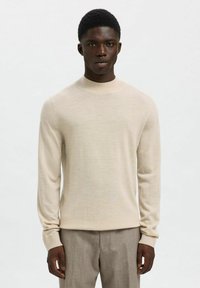 Pull en tricot beige avec un col rond et des manches longues, fabriqué dans un tissu doux, avec une texture lisse et une coupe ajustée.