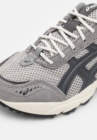 Grijze mesh en suède hardloopschoen met een gestructureerd oppervlak, witte veters en donkere accenten. Zichtbaar logo-detail op de tong.