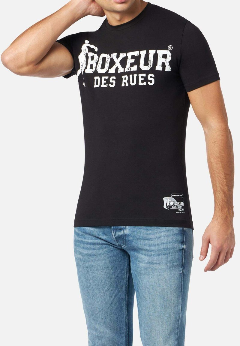 Schwarzes T-Shirt mit kurzen Ärmeln, das einen weißen Vintage-Print mit der Aufschrift "BOXEUR DES RUES" und einem Logo-Patch nahe dem Saum zeigt.