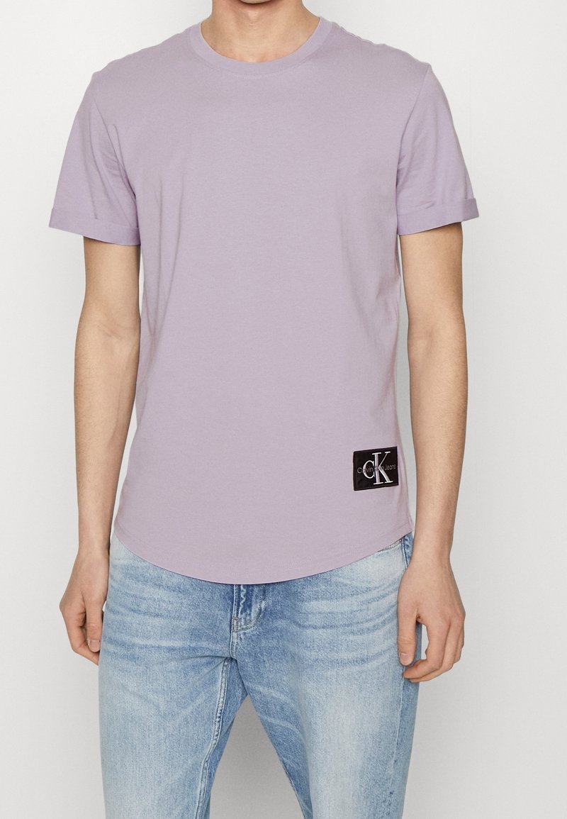 Levandulové bavlněné tričko s kulatým výstřihem, krátkými rukávy a zakřiveným lemem. Na levé straně se nachází černá čtvercová nášivka s logem.