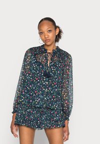 Blusa a fantasia floreale di un blu navy scuro con tessuto trasparente, orlo arricciato, maniche lunghe e fiocchetto al collo. Abbinata a una gonna arricciata coordinata.